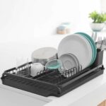 התקן ייבוש כלים גדול שחור Brabantia יושב על משטח לבן, מחזיק בצורה מסודרת צלחות, קערות, כוסות, כלים ומיכל. ירק וצנצנות מטבח מספקות תוספת סטיילינג והום דקור ברקע הפוקוס הרך. - vavaVoom.co.il