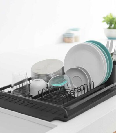 התקן ייבוש כלים גדול שחור Brabantia יושב על משטח לבן, מחזיק בצורה מסודרת צלחות, קערות, כוסות, כלים ומיכל. ירק וצנצנות מטבח מספקות תוספת סטיילינג והום דקור ברקע הפוקוס הרך. - vavaVoom.co.il